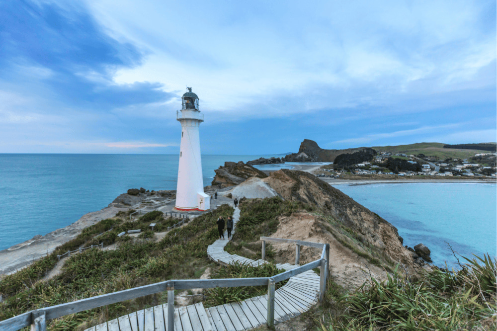 A lighthouse sits on a hill in between two bodies of blue water. There is a boardwalk leading down to the lighthouse with people walking
