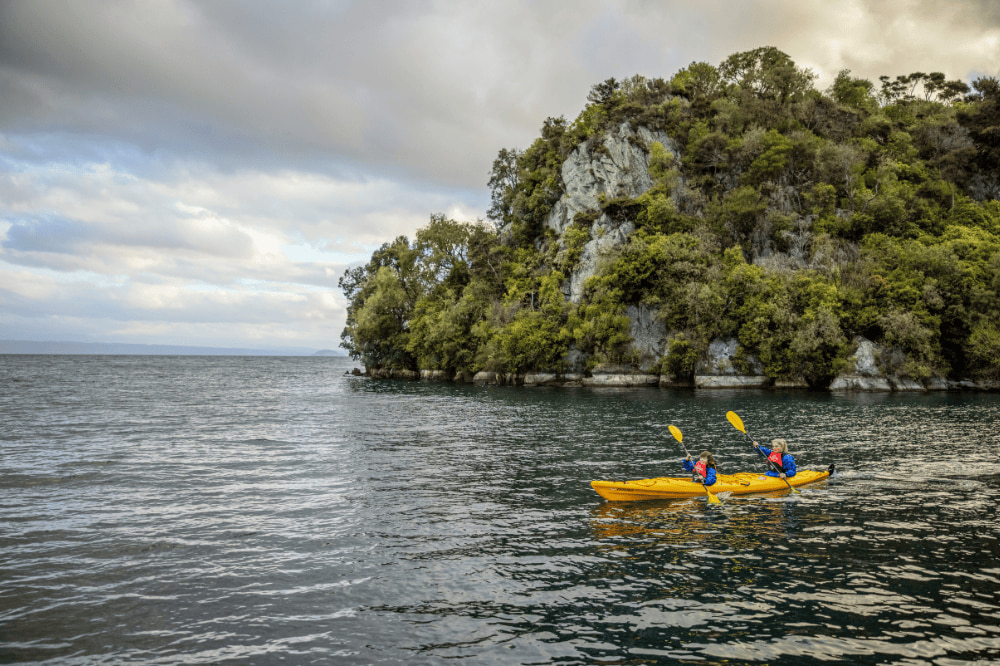 Two people kayaking on a lake with a large rock with trees behind them