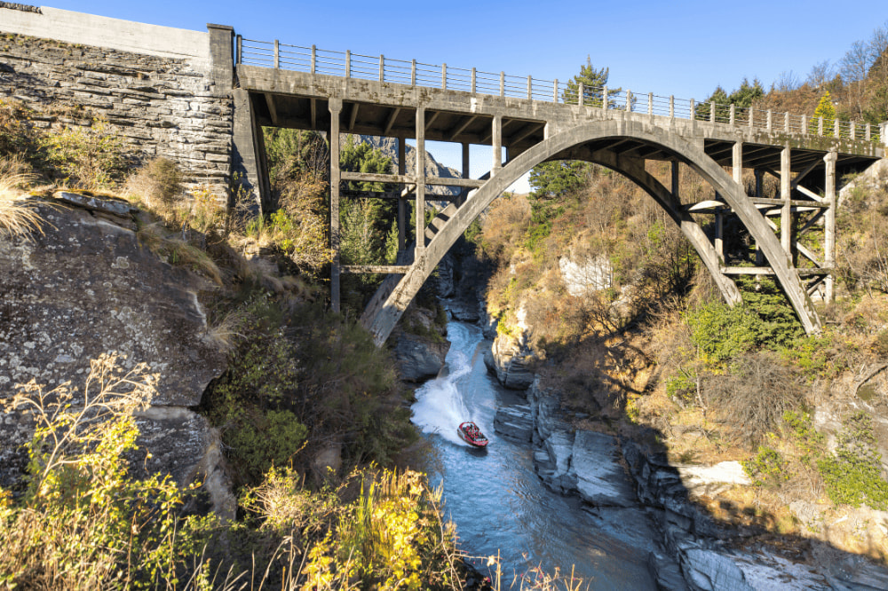 A bridge with a large arch sits high over a river. A boat is driving in the water with white water in its wake.