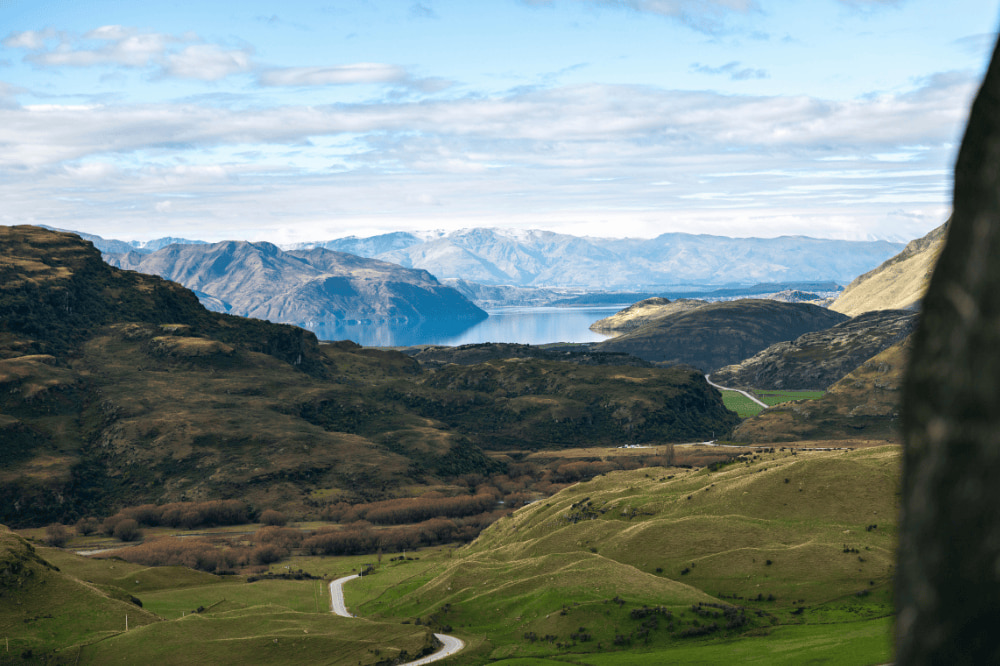 View from the top of a hill over green fields and in the distance a clear and still lake with mountains surrounding