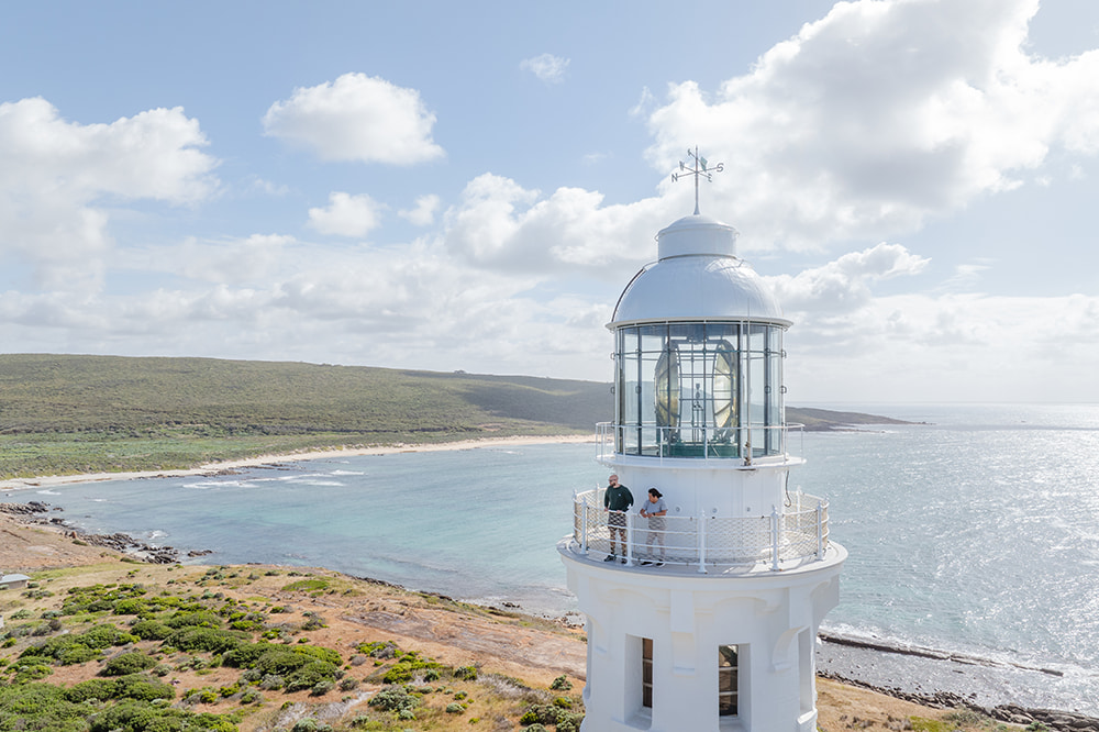 A lighthouse with two people overlooking the view. The expansive ocean is in the background