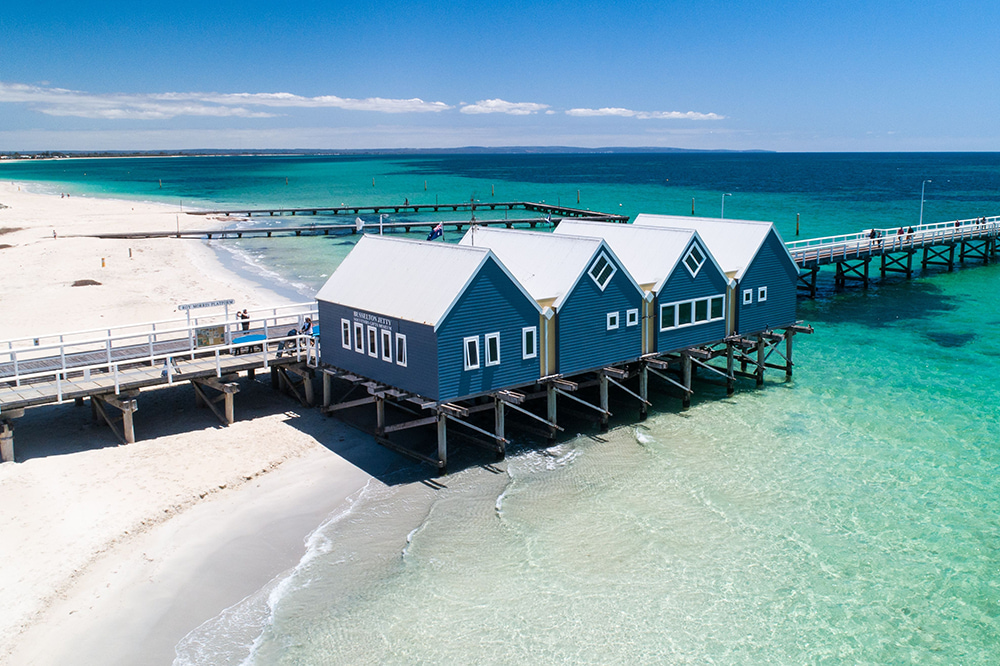 A view of Busselton Jetty from above. The water is clear and bright blue. There are four blue buildings and the jetty extends far out into the ocean