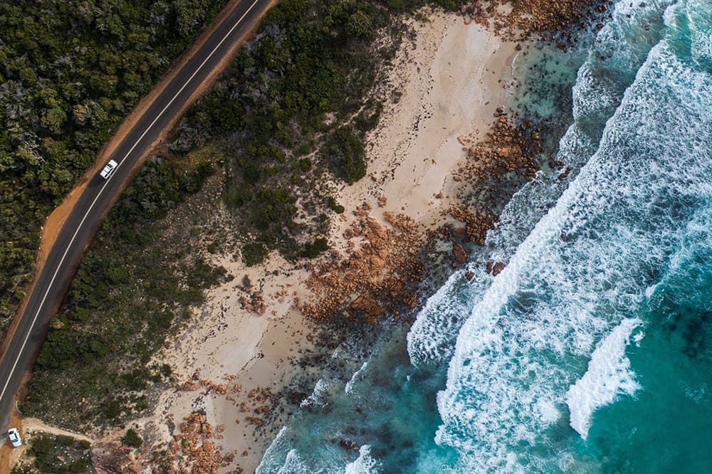 A car drives along a coastal road. There are trees on either side of the road and a rocky, wavy beach along side.