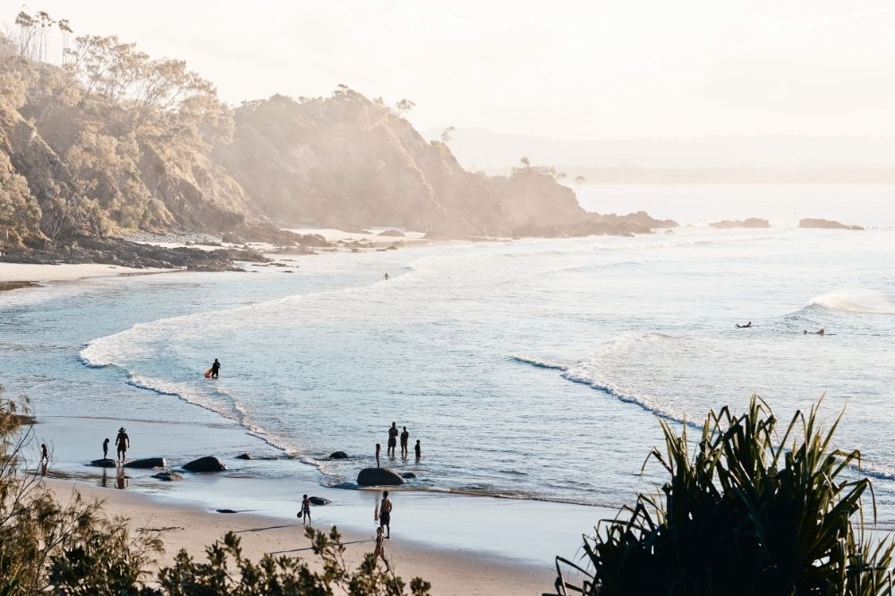 The view of a beach from above. Trees and rocks surround the beach while people swim and walk along the shore.