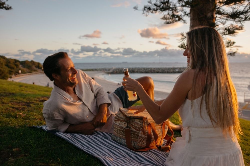 A couple sit on a picnic rug at sunset with the beach in the background