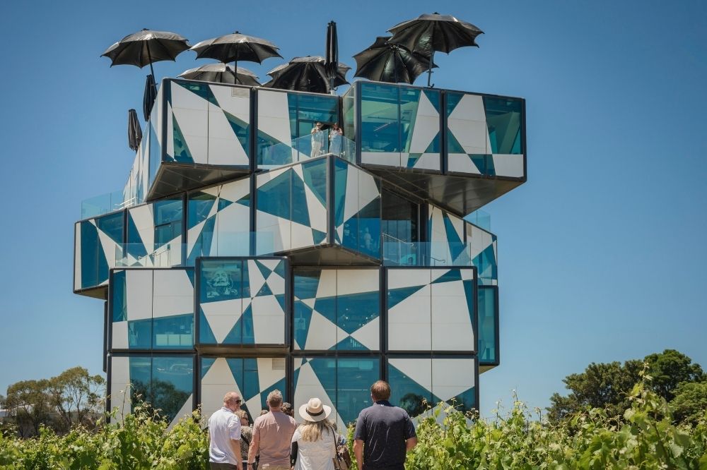 A group of people look up at a building shaped like a rubrics cube. Three are black umbrellas open on the roof