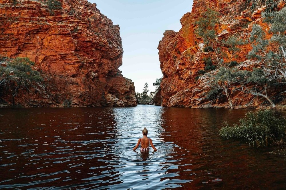 A woman in striped bathers stands waist deep in a creek. She looks out at two large red rock formations. There are trees on the edges of the creek