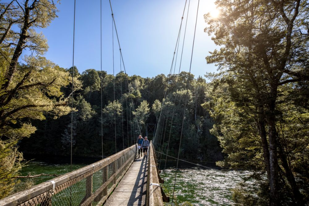 Two people walk along a large suspension bridge, the water below is running quickly. Large green trees surround.