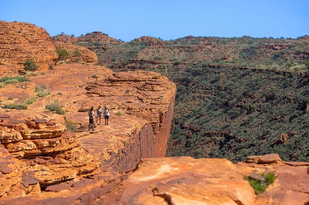 A group of people stand on a large red rock formation looking out at a vast canyon lined with trees