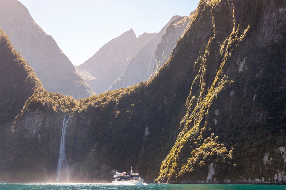 A boat floats in the water, looking small compared to the tall, green rock formations surrounding. A waterfall cascades down the rocks in the background.