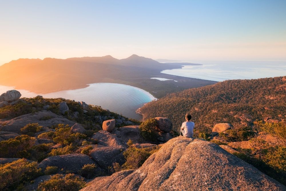 A person sits on a rock high above a bay. Clear blue water and white sand is visible below