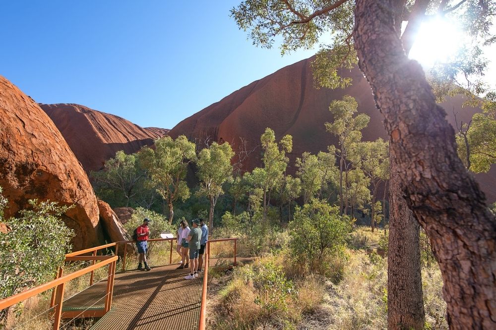 A tour group stands on a walking platform looking out at Uluru in the background