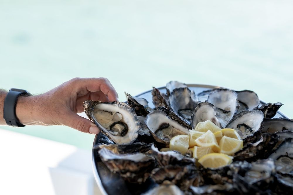 A person grabs an oyster from a plate stacked with oysters and fresh lemon wedges.