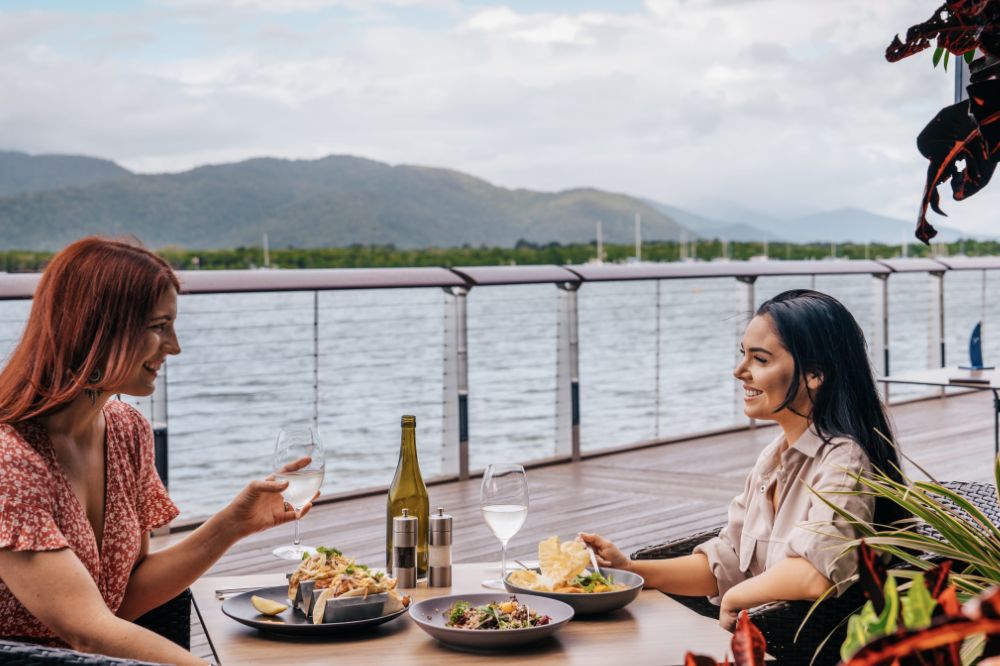 Two people dine outside by the waterfront. With glasses of wine and large plates of food