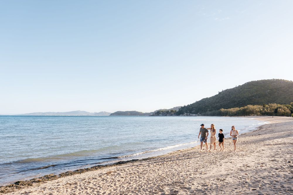 A family walk along a secluded beach with white sand and blue water. There are tree lined hills in the background