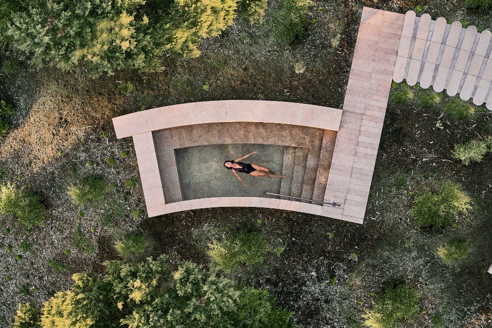A woman lies flat in a stone hot spring. The surrounds are quiet with native plants