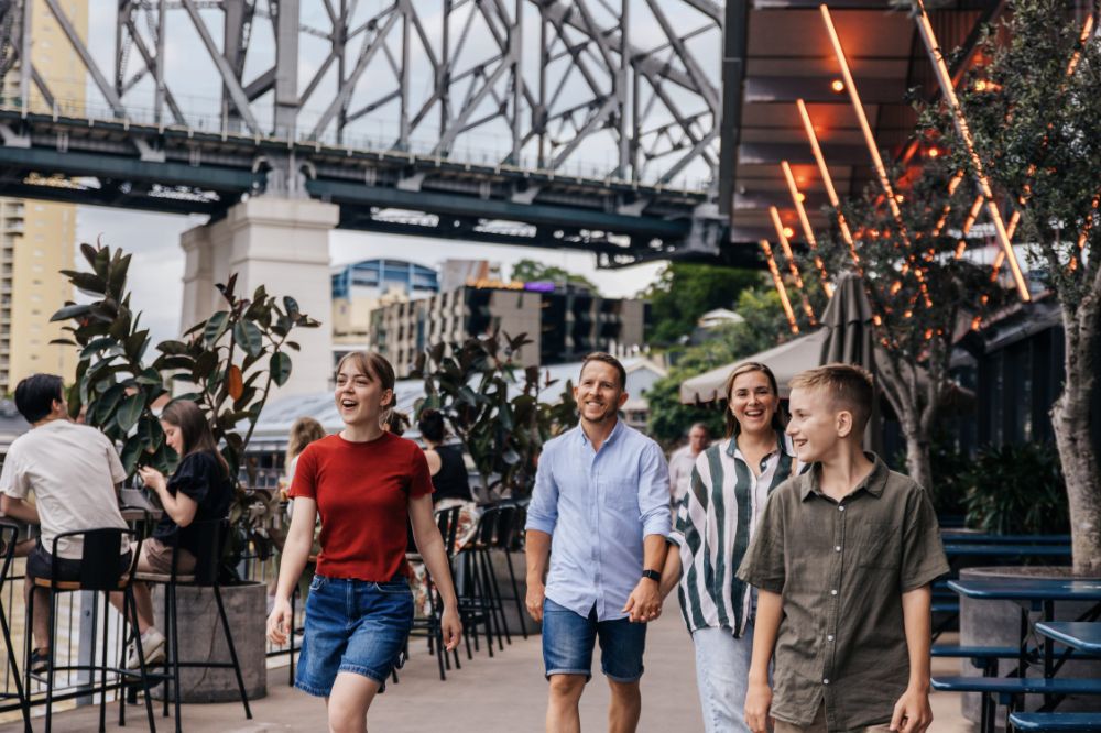 A family walk along a wharf with a large bride above. There are people sitting at the tables of a restaurant overlooking the river.