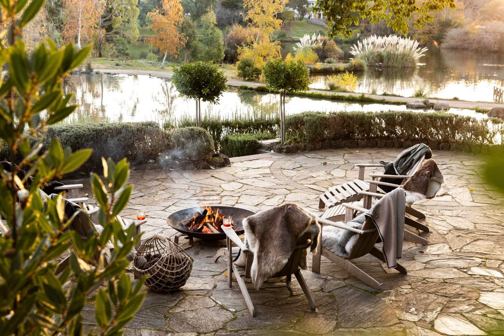 A group of chairs are huddled around a fire pit, with a tidy garden and a lake in the background