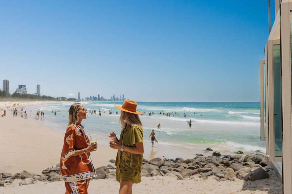 Two women stand in front of the beach with iced coffees in hand. There are people and surfers in the water.