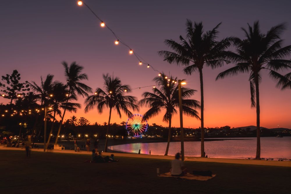 Palm trees and a beach at sunset. The sky glows orange and purple with a lit up ferris wheel in the background