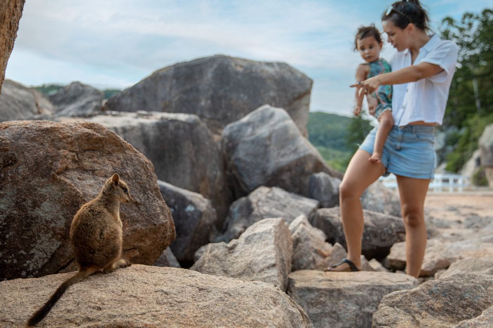 A woman holds a child and points at a small animal sitting on a rock