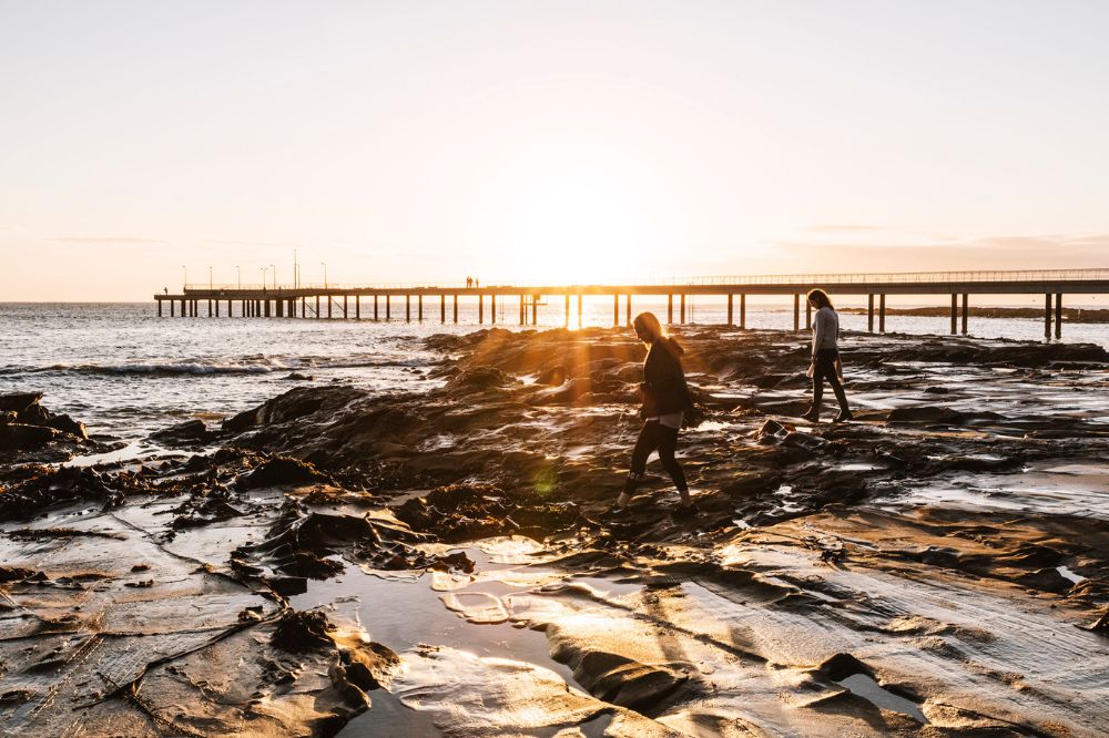 Two people walk along the rocky foreshore with a long jetty in the background. The sun sets with orange brightness.