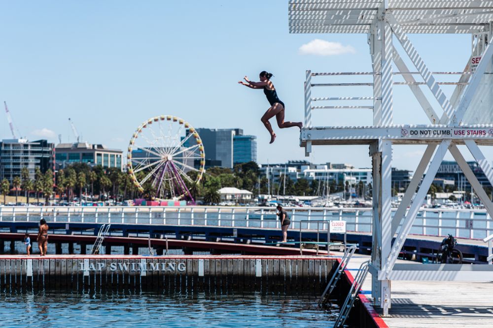 A person jumps off a diving platform into the ocean. There are sailboats and a ferris wheel in the background