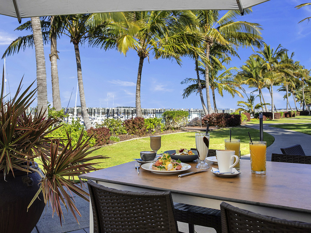 A hotel breakfast on a table with grass and palm trees surrounding. There are boats in the marina in the background