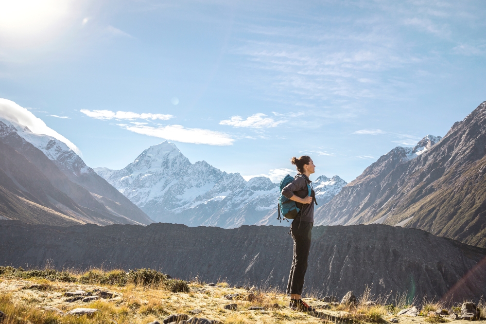 A person with a backpack looks out at the surrounding mountains with snow capped peaks