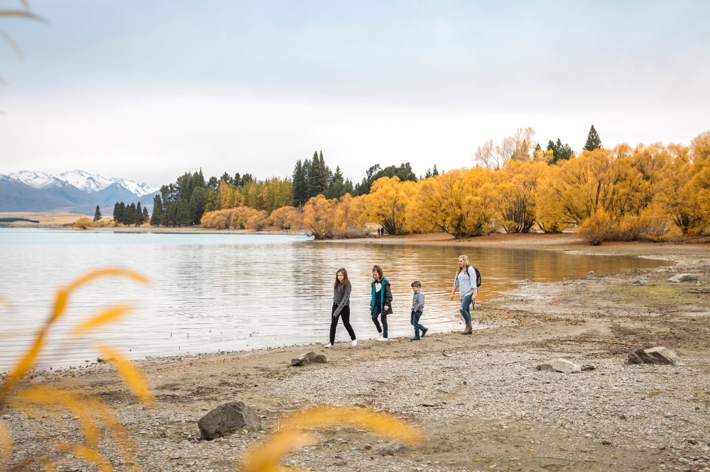 A family walk along the shore of a lake that is surrounded by yellow autumn trees and snow capped mountains in the background