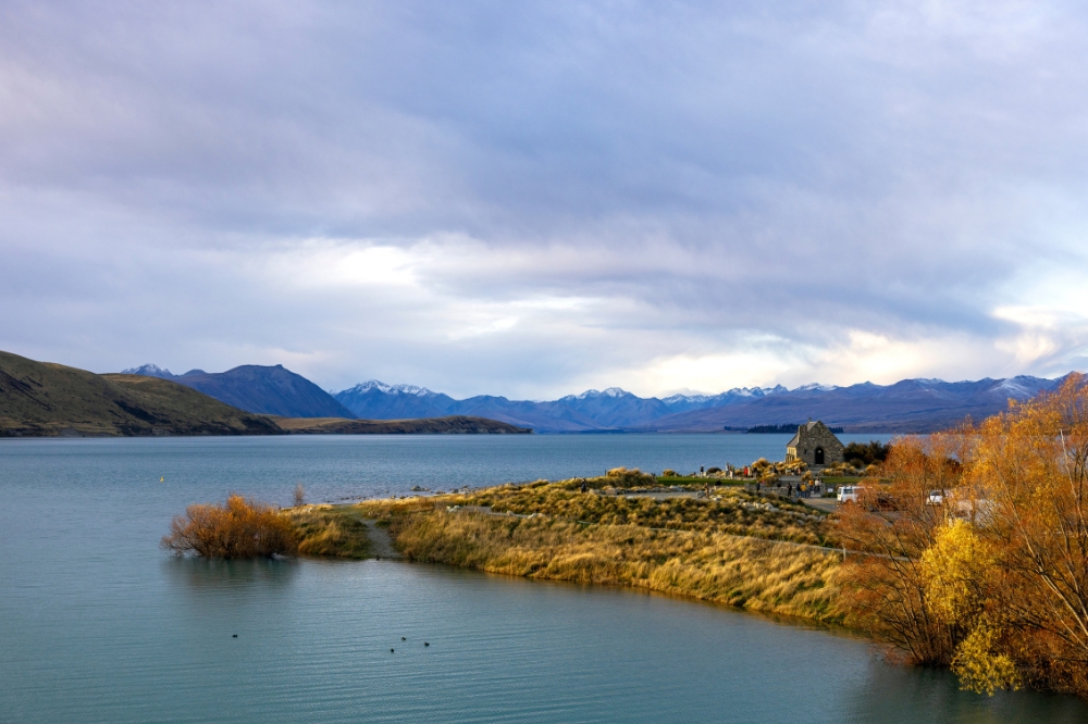 A stone church sits on the edge of a large lake. There are autumn coloured trees along the edges and snow capped mountains in the background