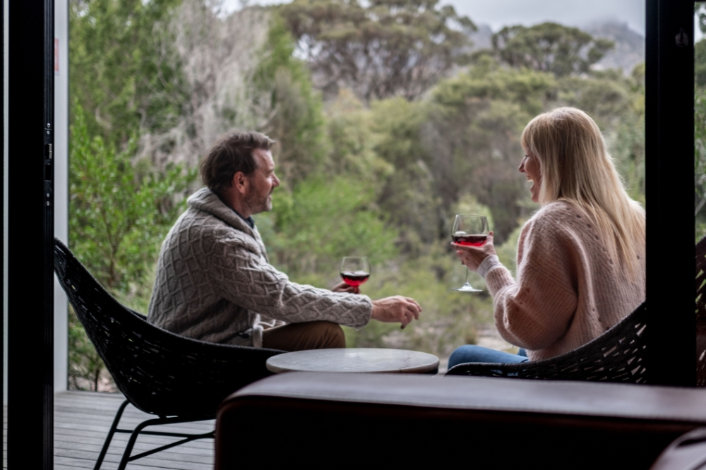 Two people sit on a deck surrounded by bushland, smiling and holding glasses of red wine