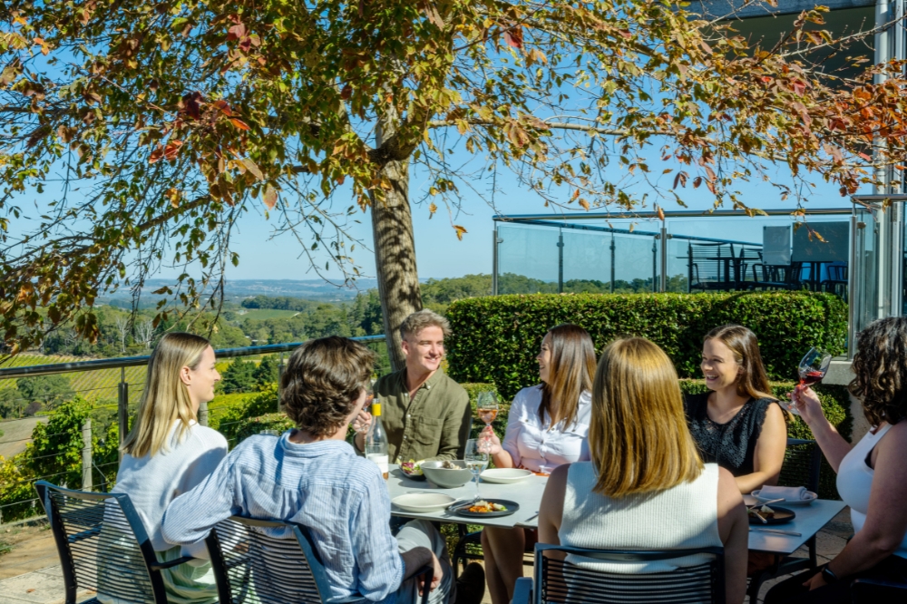 A group of people sit around a table with glasses of wine. An autumn tree hangs above them