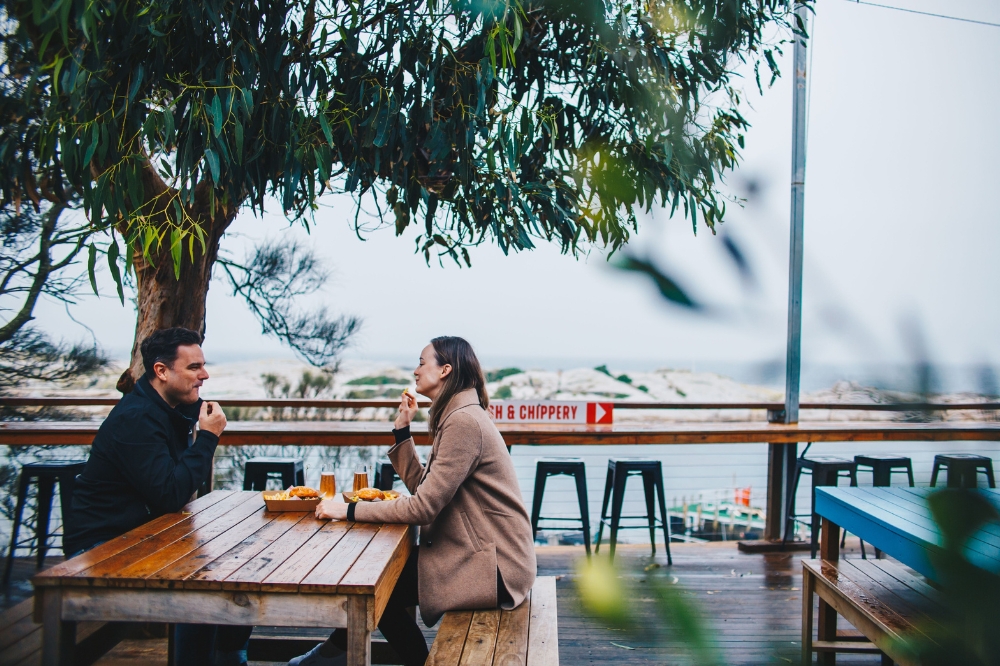 People dine on an oceanside deck under a gum tree