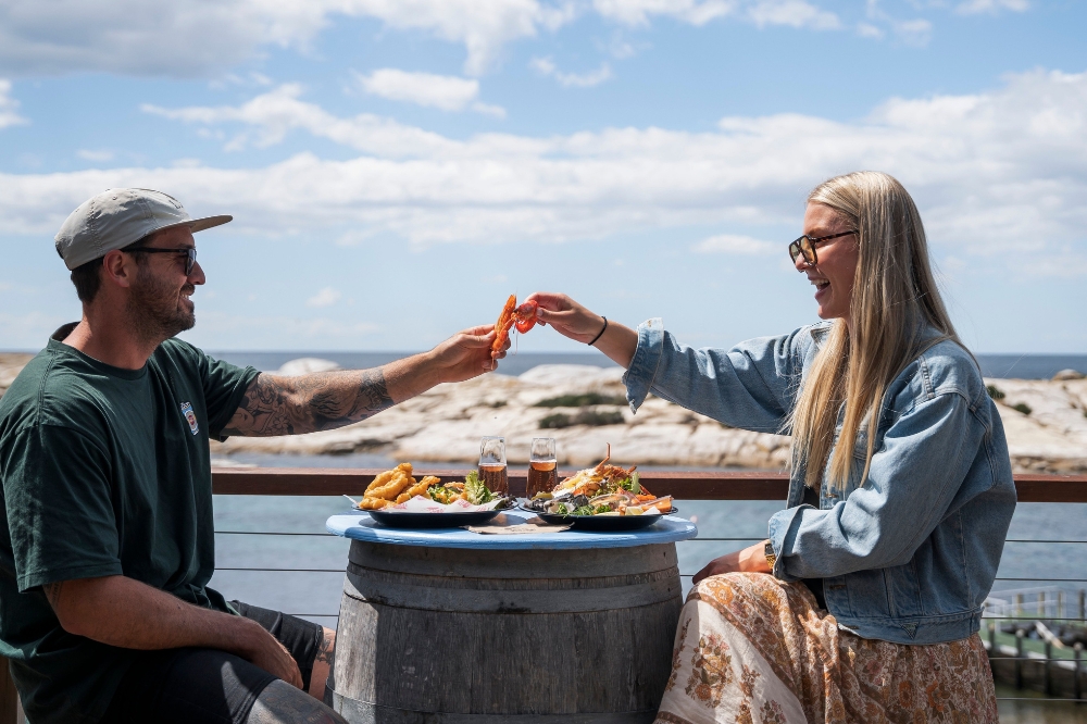 Two people dine on an oceanside deck. They are holding seafood in their hands and touching it together