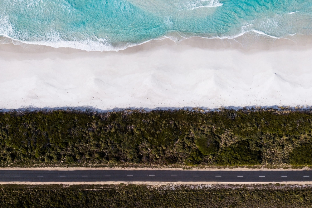 A straight road with green trees and a beach with white sand a clear blue water