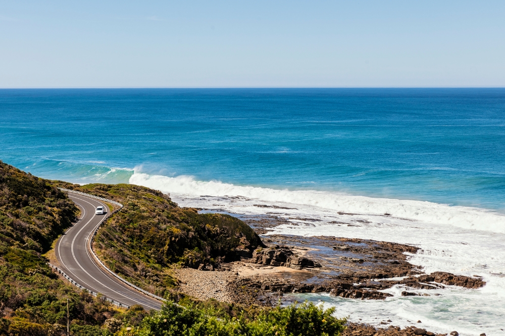 A car drives along a winding coastal road. The ocean forms large surf waves