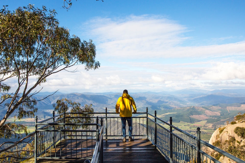 A person in a yellow jacket stands at a lookout overlooking hills and mountains
