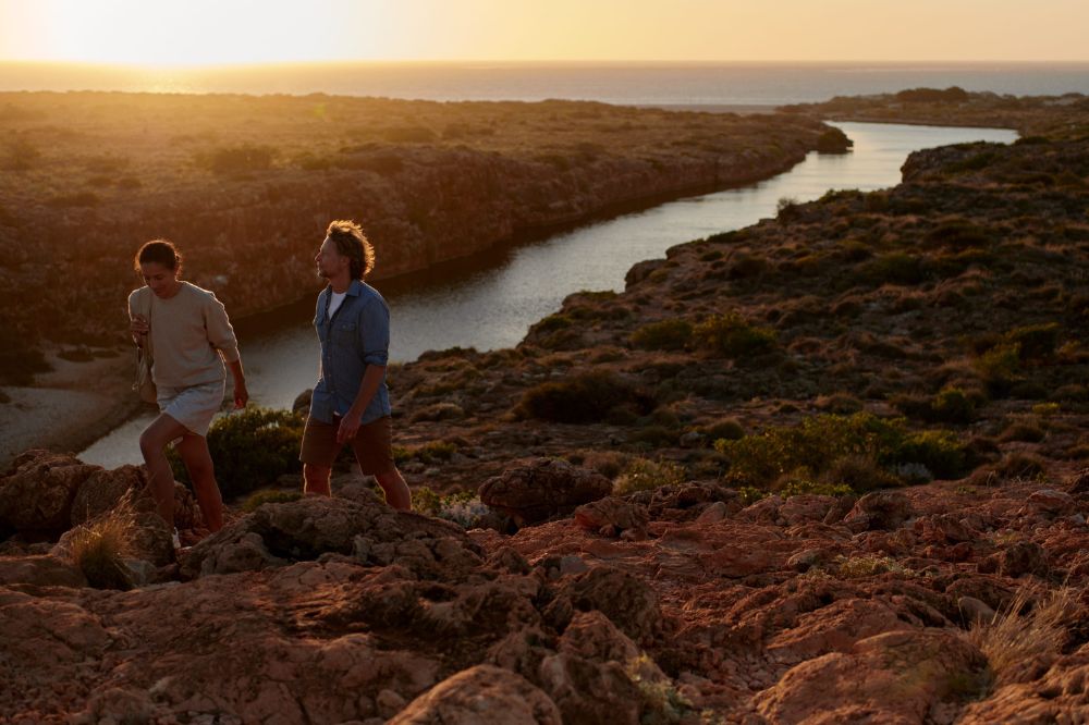 Two people walk along a rocky walking track above a river mouth meeting the ocean