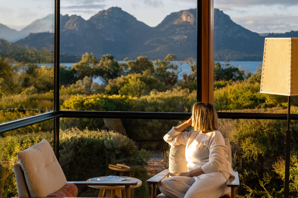 A person looks out a window at trees, water and rocky mountains in the background