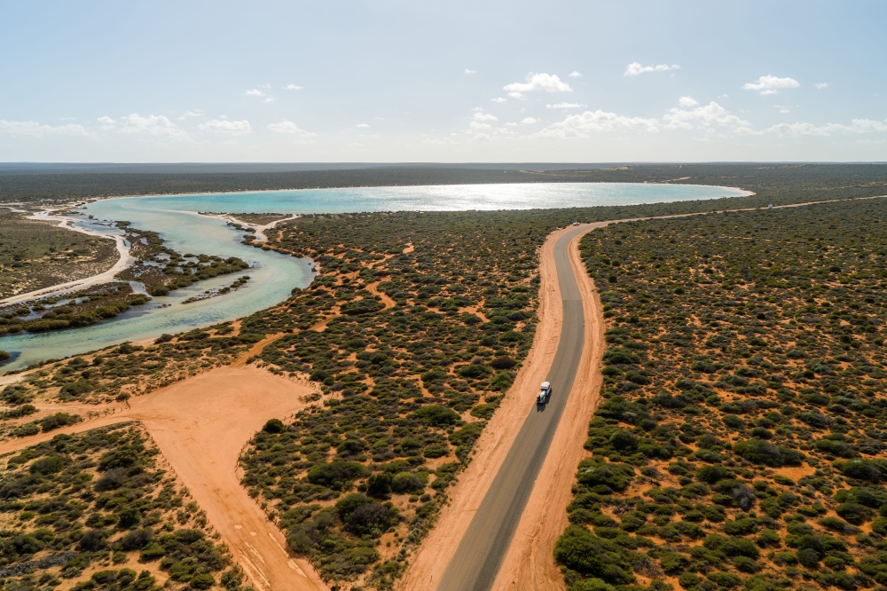 A car drives down a country road with red dirt, green bushes and blue water